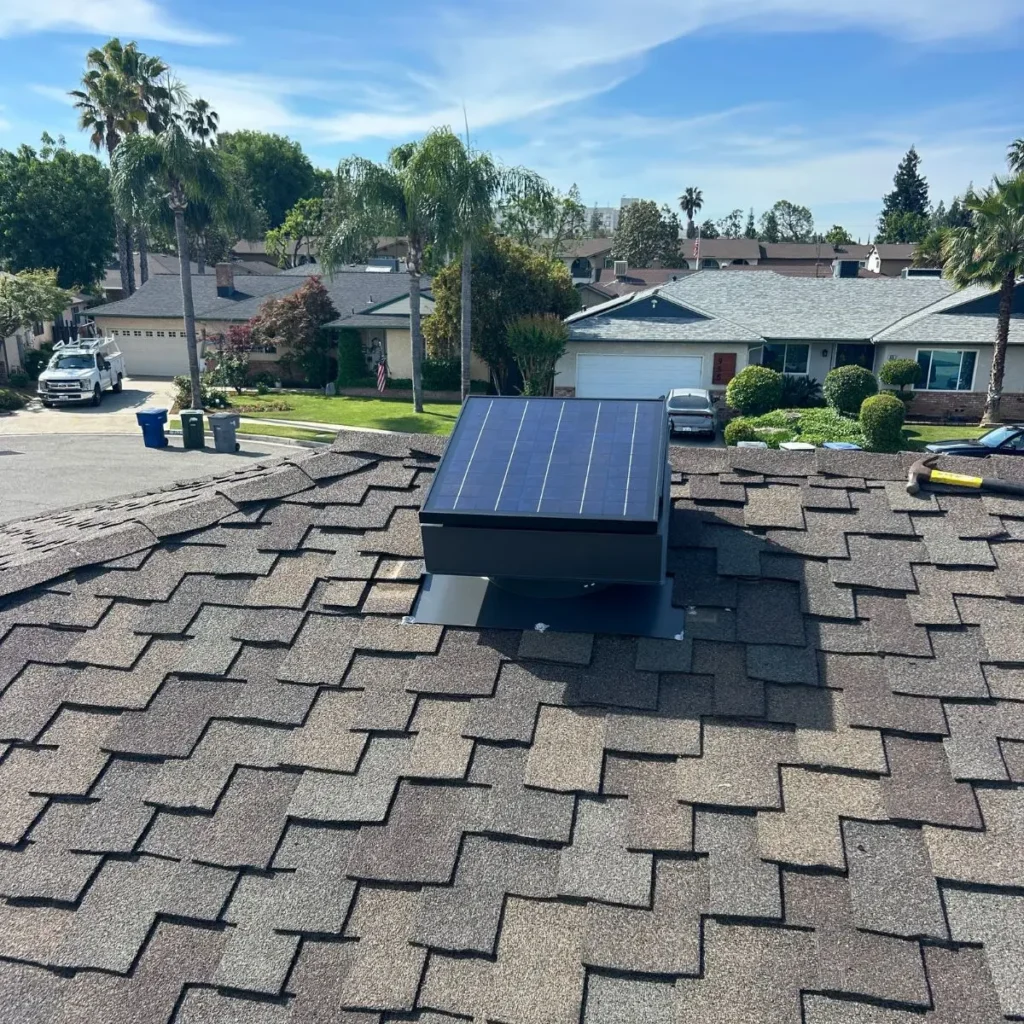 A solar panel is mounted on a roof with brown shingles in a suburban neighborhood. Palm trees and houses with green yards are visible under a blue sky.