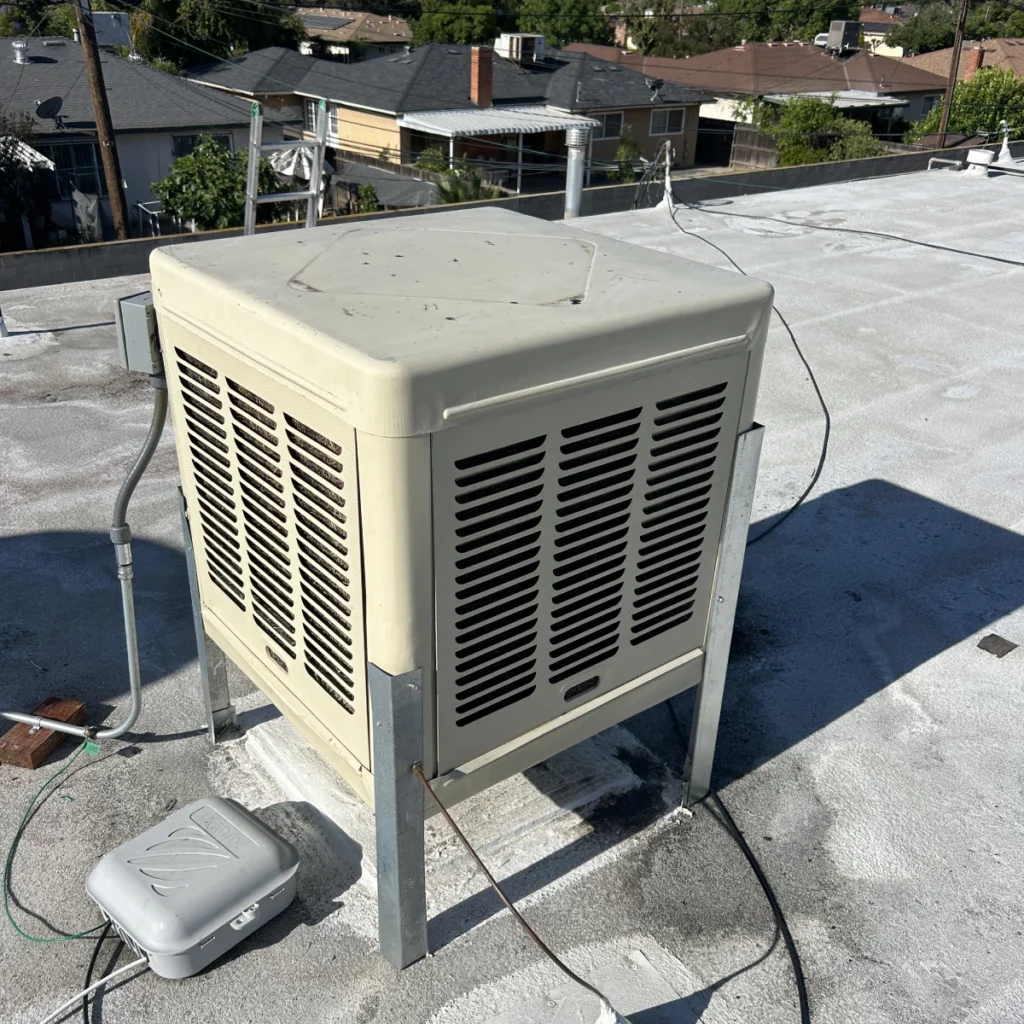 A beige rooftop HVAC unit stands on metal legs, surrounded by wires and pipes, with neighboring houses and trees visible in the background under a clear sky.