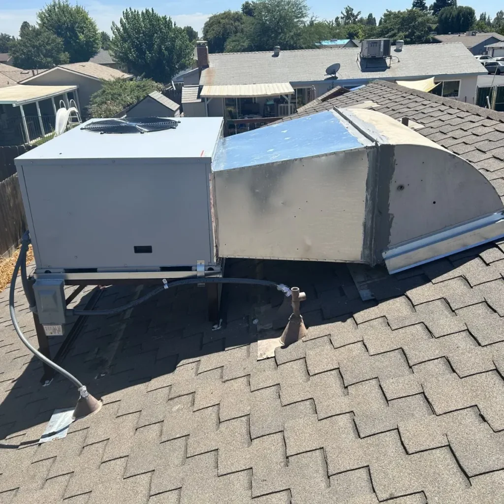 Rooftop view of an HVAC unit connected to a duct on a shingled roof, with nearby vent pipes and residential houses visible in the background under a clear sky.