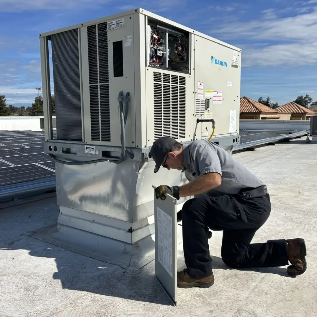 A technician kneels on a rooftop while repairing an air conditioning unit. He is holding an open panel, and solar panels are visible nearby under a partly cloudy sky.