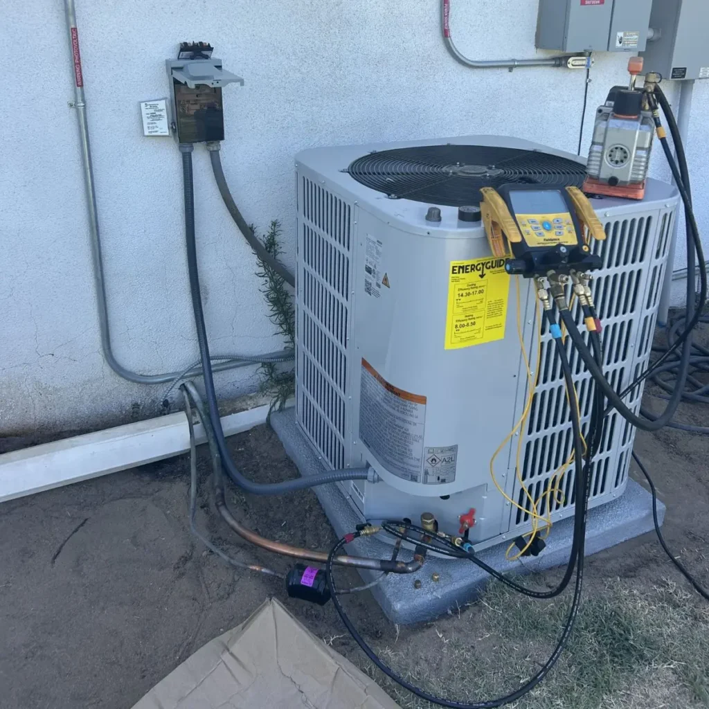 A central air conditioning unit is being serviced outdoors. Gauges and hoses are attached to the unit, and tools are nearby. The unit sits on a concrete pad next to a white wall with electrical conduits above it.