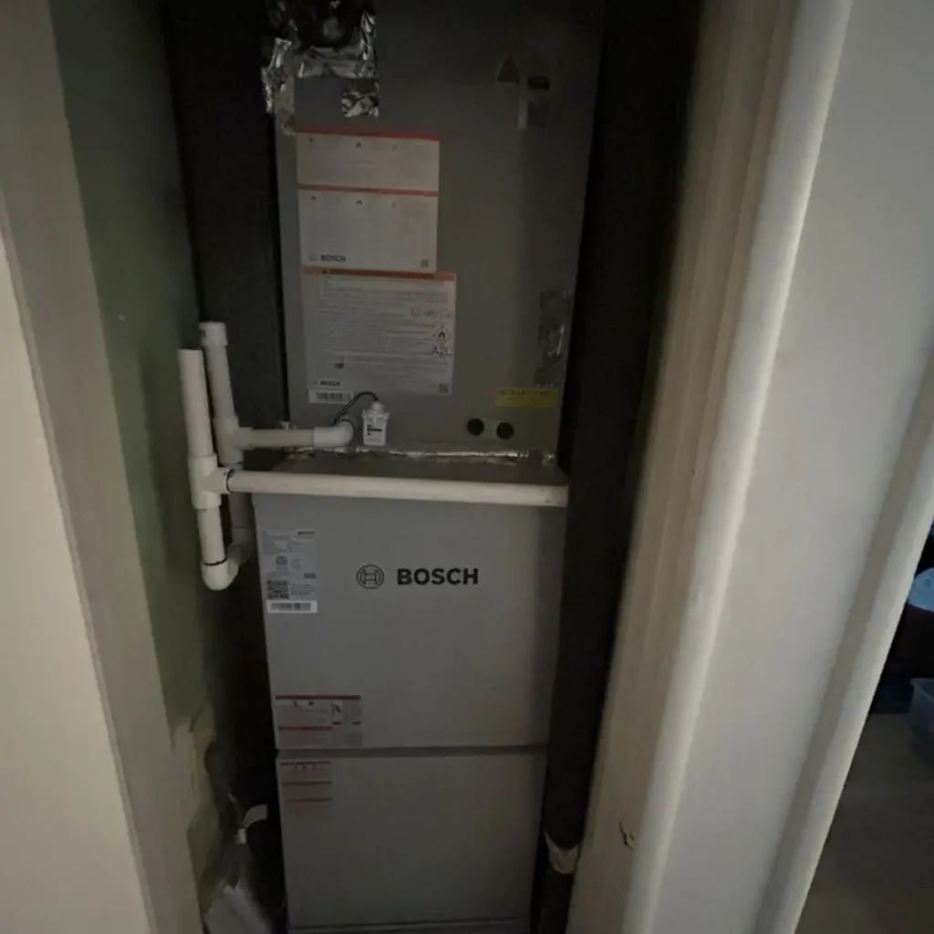A Bosch HVAC unit installed in a small, narrow closet with visible white PVC pipes, electrical wiring, and instruction labels on the unit.