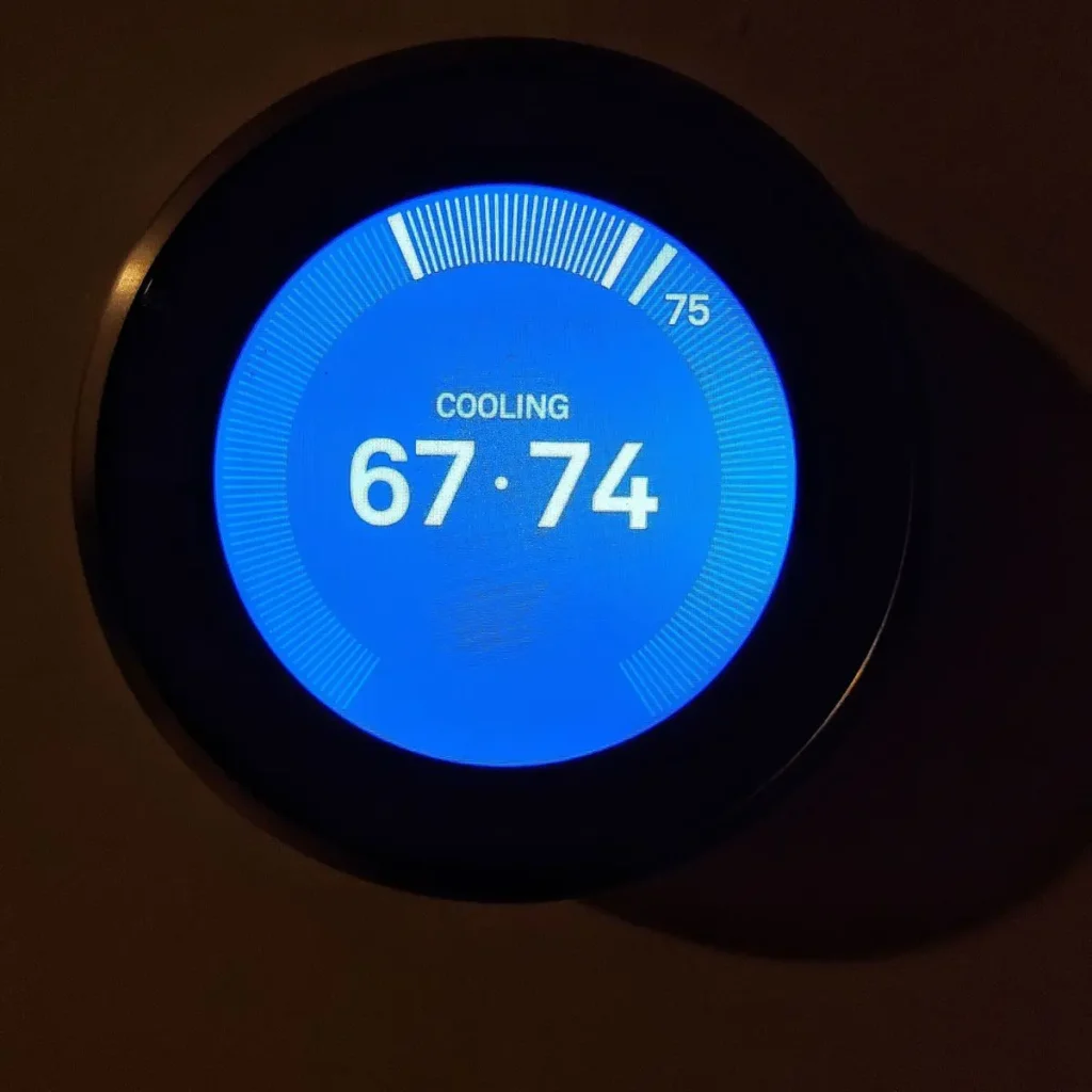 A digital thermostat displays a cooling mode with a set temperature of 67 degrees and a current temperature of 74 degrees, shown on a bright blue circular screen.