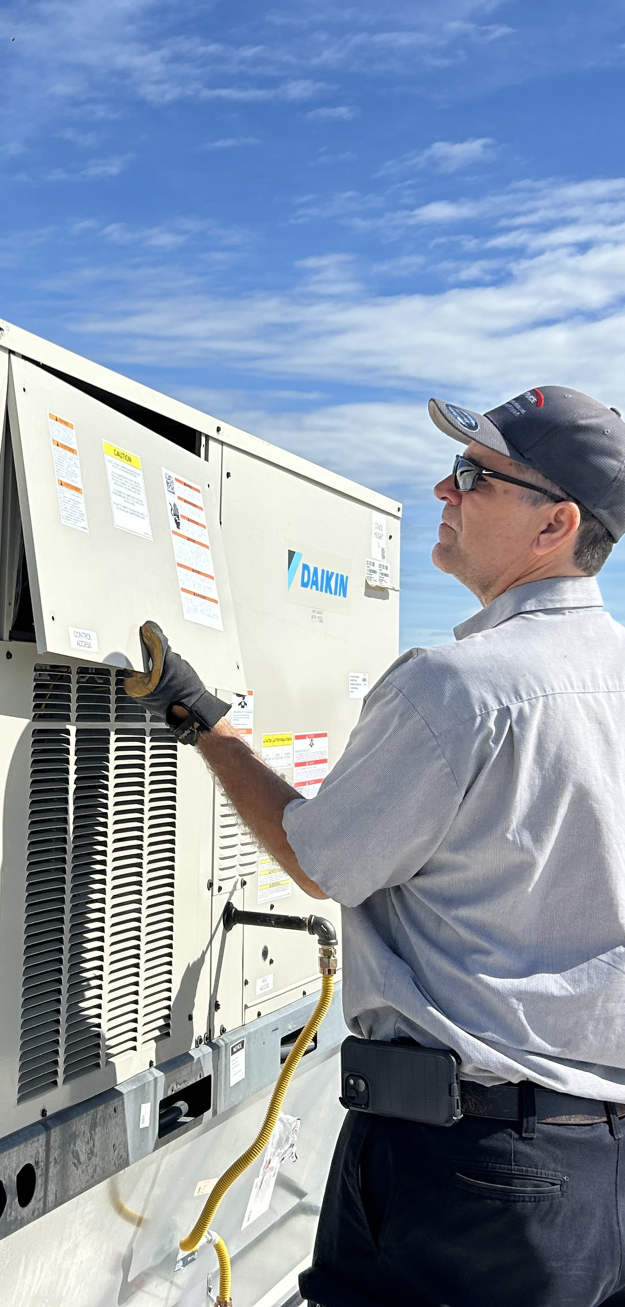 A technician in a gray shirt and cap inspects a Daikin HVAC unit on a rooftop under a blue sky, wearing sunglasses and gloves, with tools attached to his belt.
