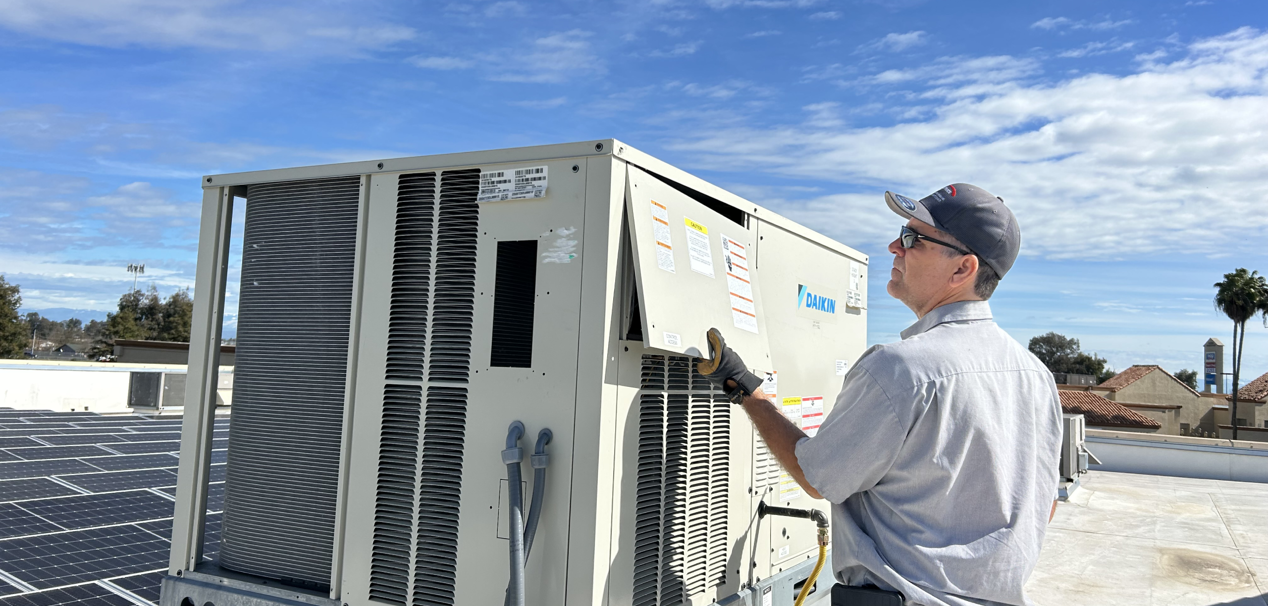 A technician wearing a cap and sunglasses inspects a rooftop HVAC unit on a sunny day, with solar panels and buildings visible in the background.