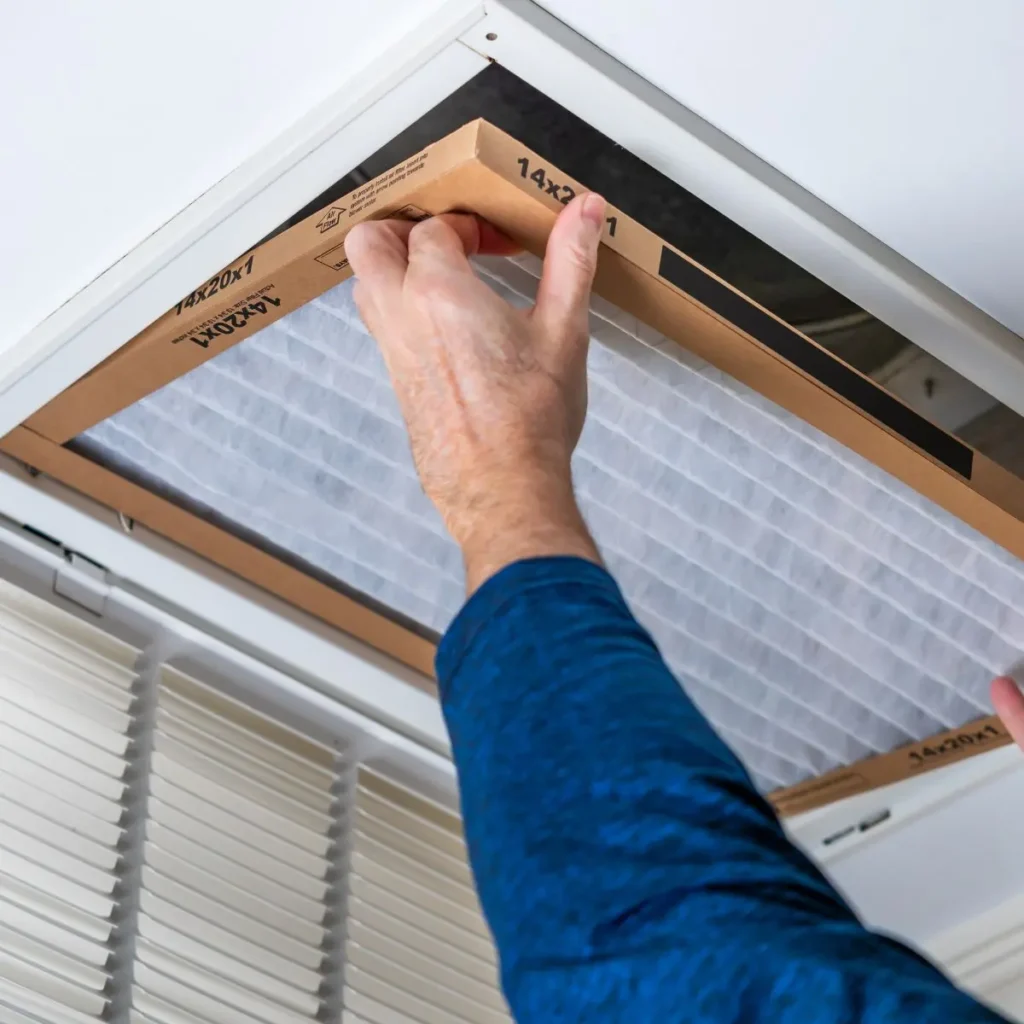 A person in a blue shirt installs a new air filter into a ceiling vent.