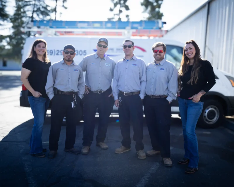 Six people, four wearing matching gray work shirts and two in black tops, stand and smile outside in front of a white van and building. The group appears relaxed, with sunlight casting soft shadows on the pavement.