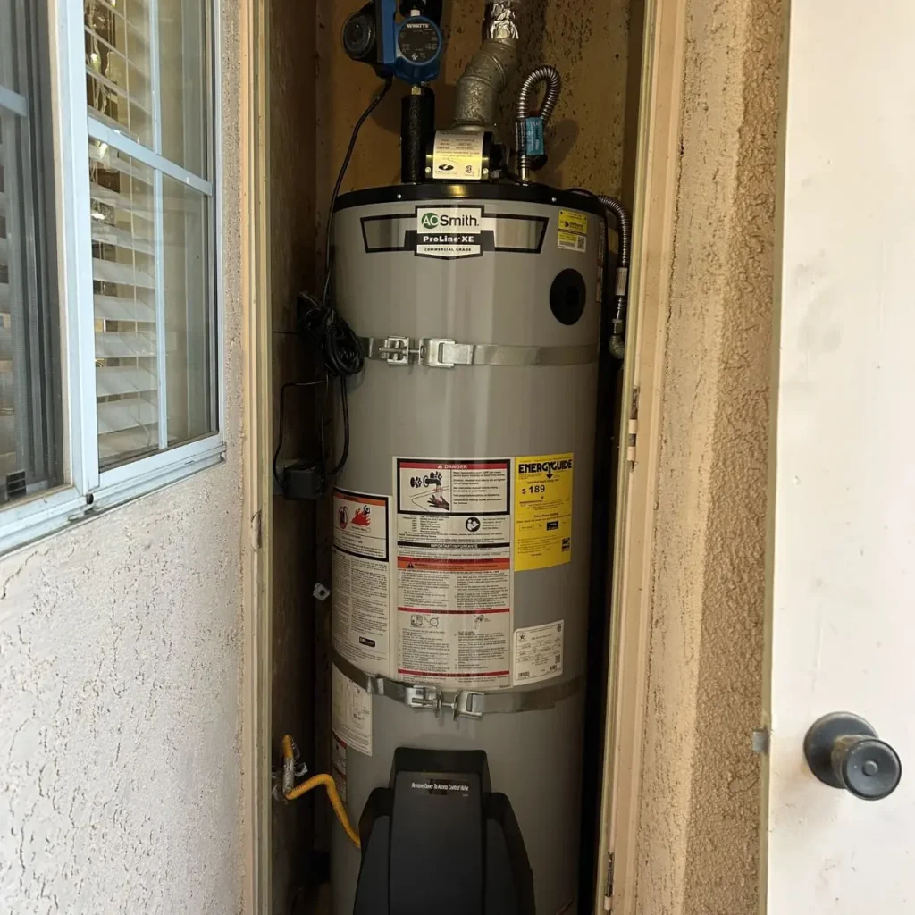 A modern water heater installed in a small, outdoor utility closet with beige walls; the unit has several labels and warning stickers, pipes, and a vent connected at the top.