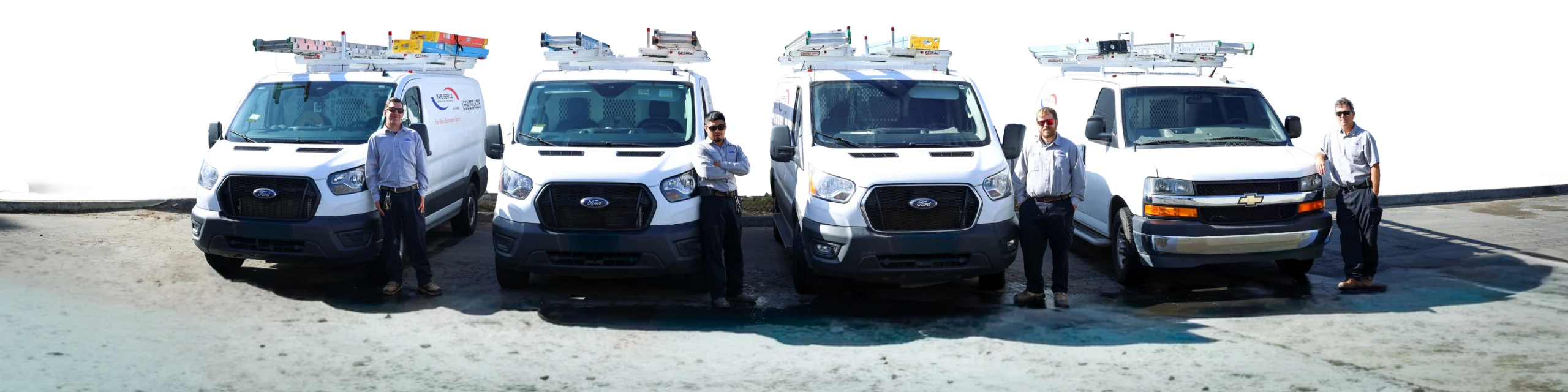 Four men in uniforms stand next to four white service vans with ladders on their roofs, parked side by side on a concrete surface against a white background.