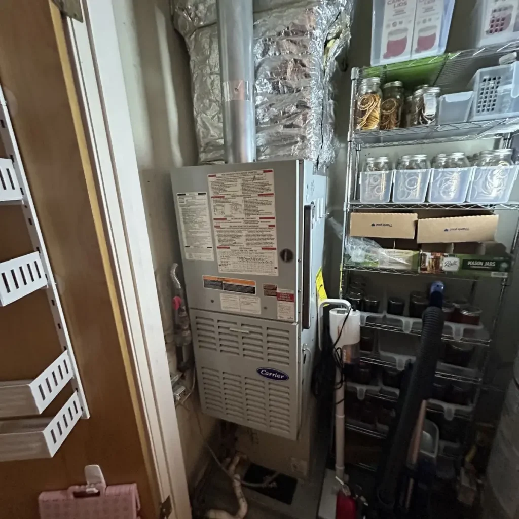 A furnace unit in a small storage room, surrounded by shelves holding jars, boxes, and household items. A vacuum cleaner is on the floor nearby. The space appears cluttered and tight.