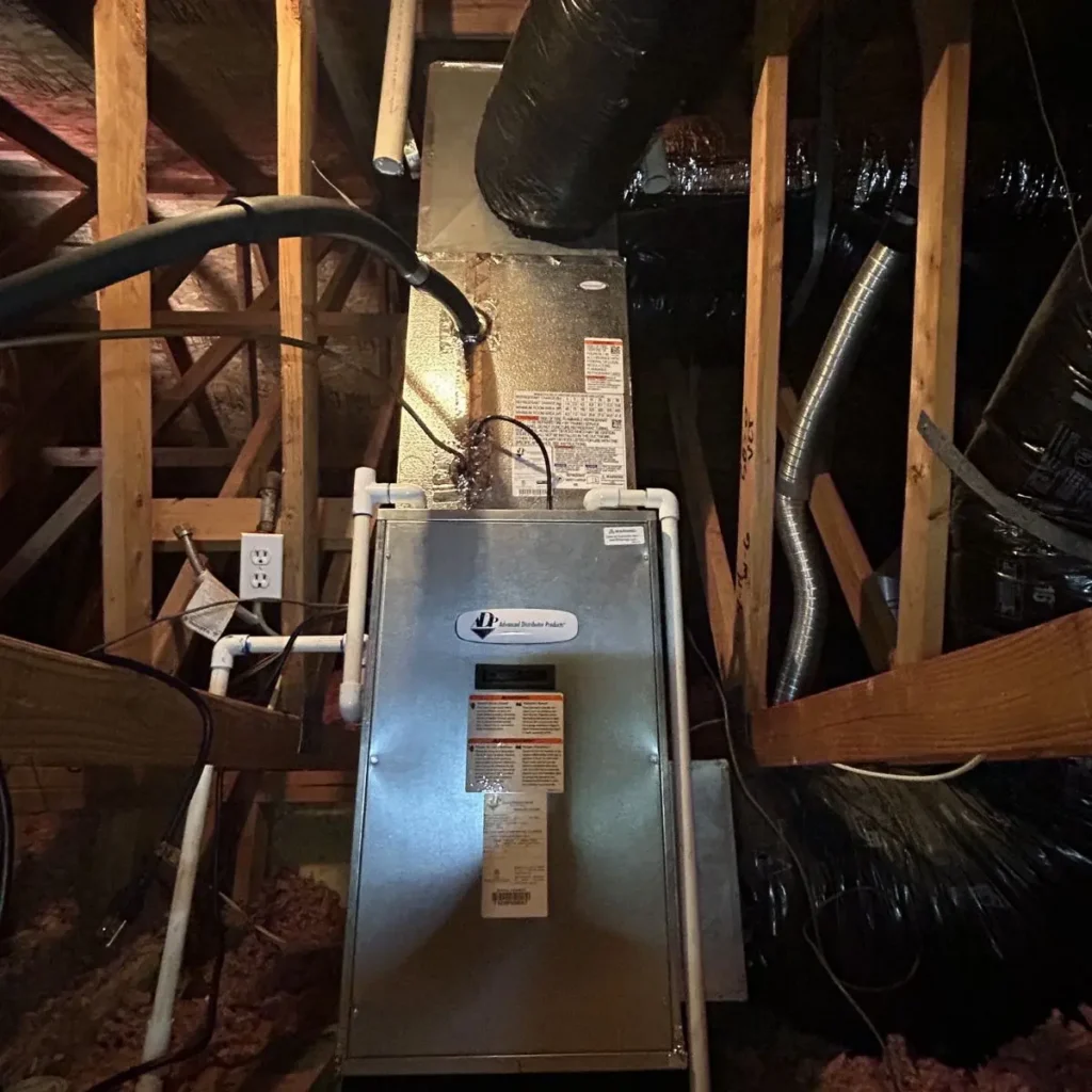 A metal HVAC unit is installed in an attic, surrounded by wooden beams, insulation, black air ducts, and piping. An electrical outlet and cables are visible on the left side of the unit.