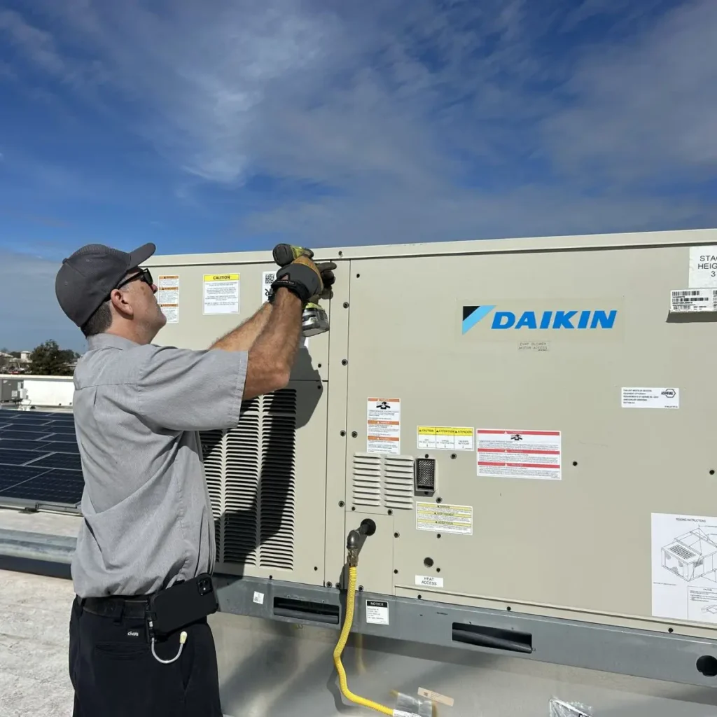 A technician in a gray uniform uses a power drill to work on a Daikin HVAC unit on a rooftop under a blue sky.