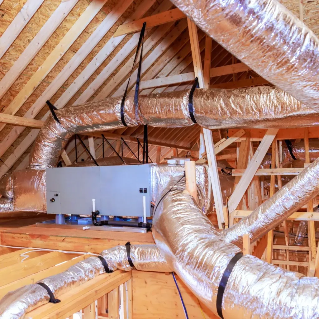 An HVAC unit with shiny insulated air ducts installed in a wooden attic, surrounded by beams and rafters.