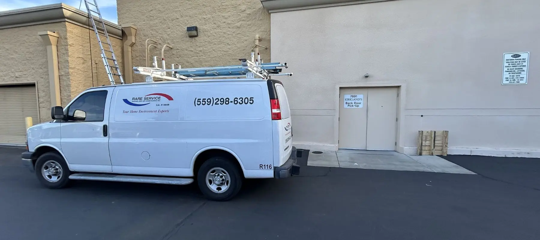 A white van with ladders on top is parked beside a beige building near a Do Not Block Door sign. The van displays a business logo and phone number on its side.