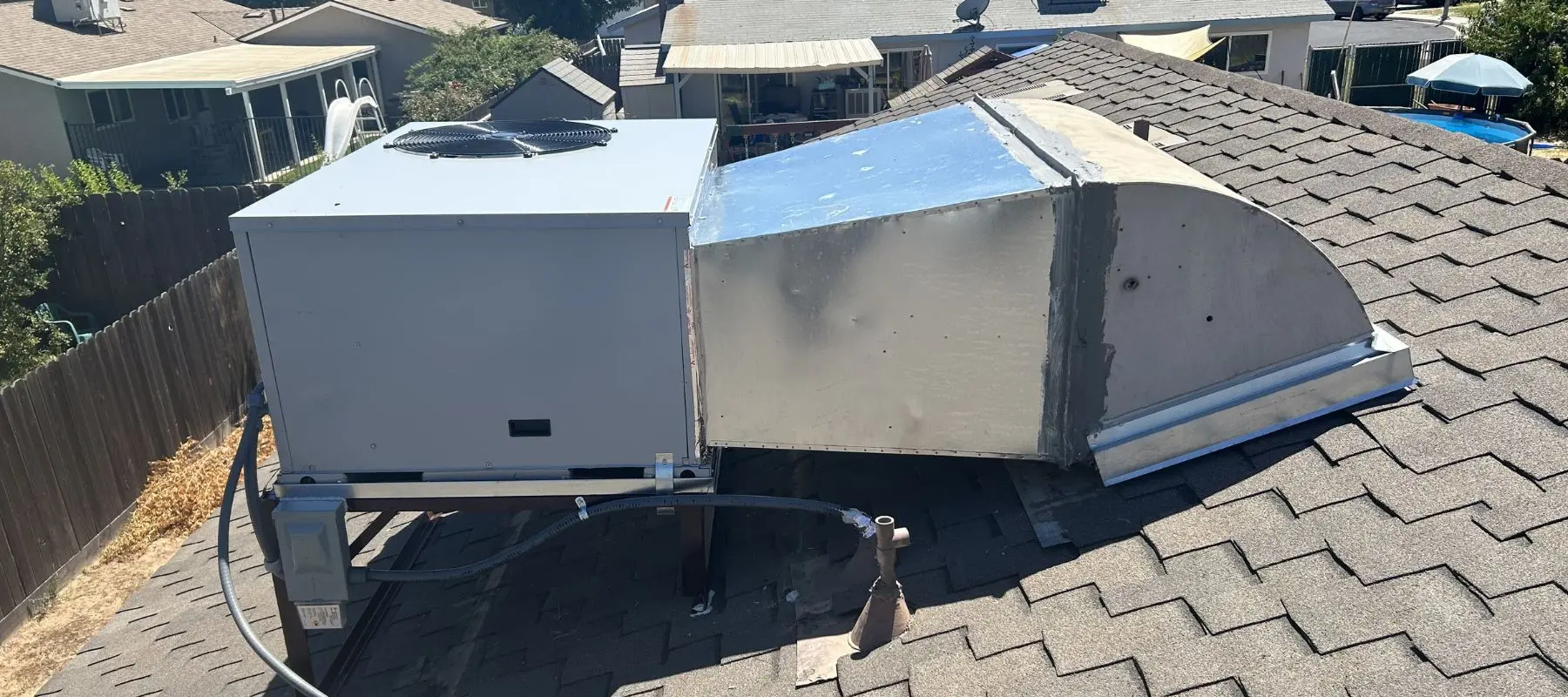 A large HVAC unit with ductwork is installed on the sloped roof of a house, surrounded by neighboring houses under sunny skies.