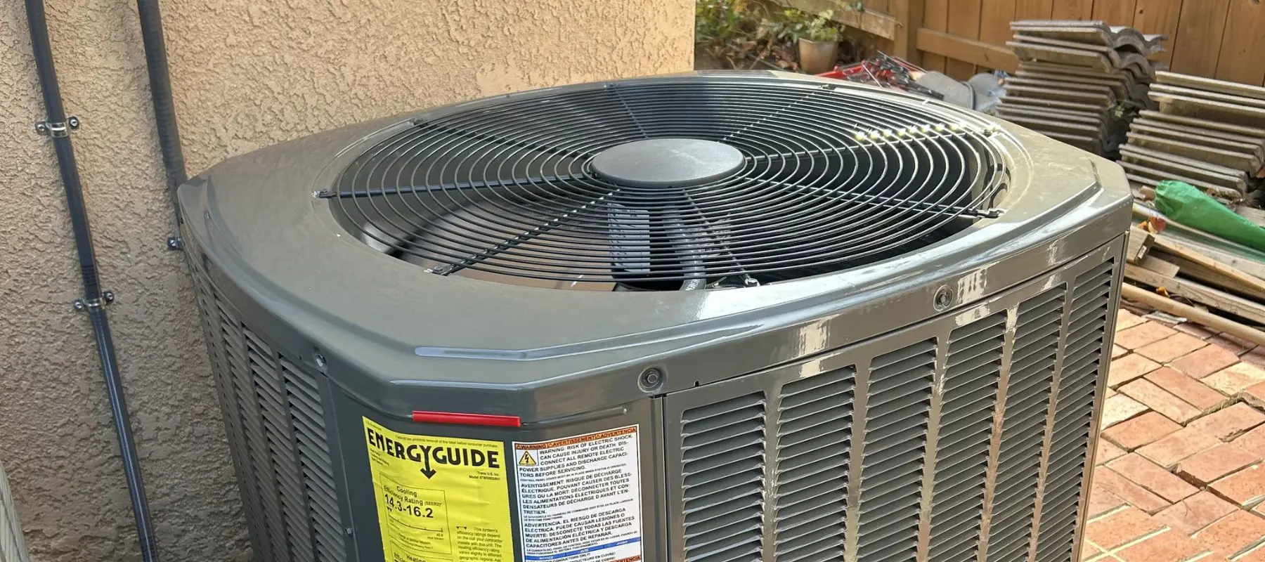 A close-up of a gray outdoor air conditioning unit with a visible fan on top, yellow energy guide label, and a brick patio in the background.