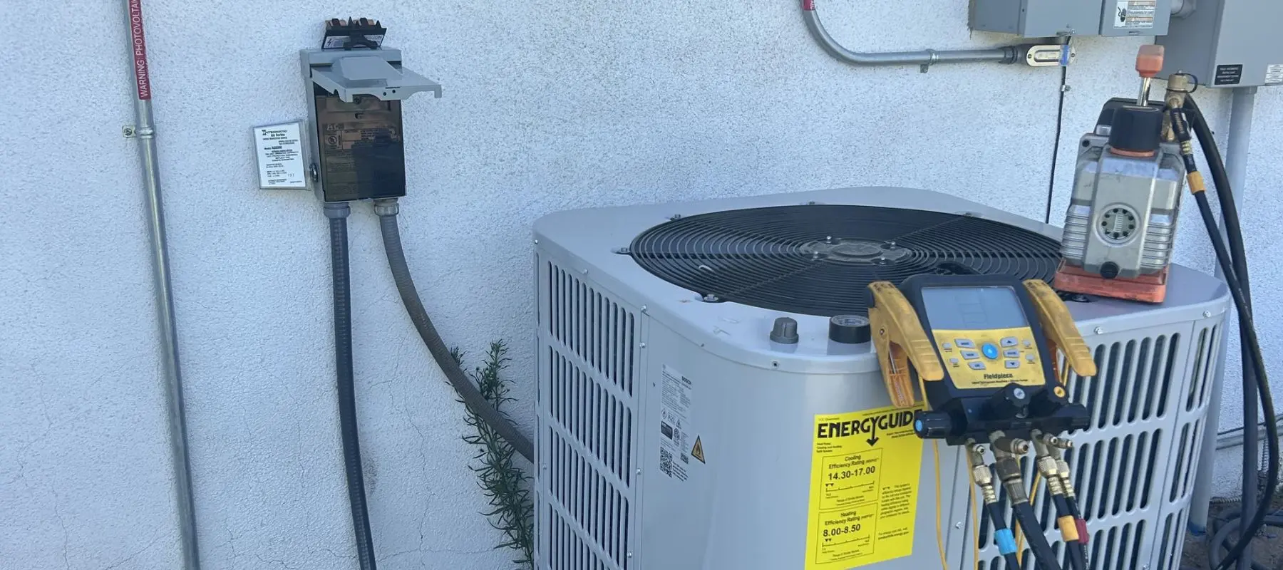 Digital gauges are attached to a central air conditioning unit outside a building, with wires and hoses connected, indicating it is being serviced or tested.