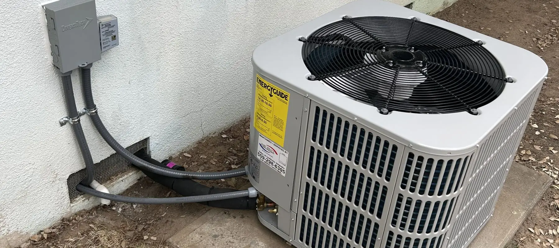 A gray outdoor air conditioning unit sits on a concrete pad next to a white wall, with connecting pipes and electrical lines running into the wall. The fan on top of the unit is visible.