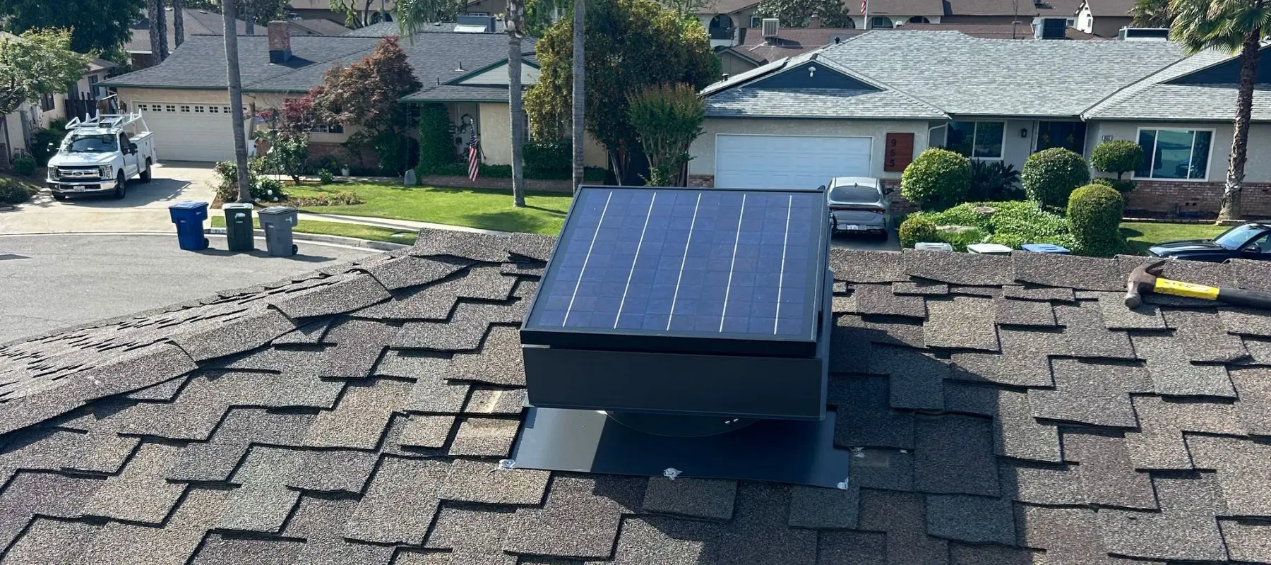 A small solar panel is installed on a rooftop with brown shingles in a suburban neighborhood; houses, cars, and greenery are visible in the background.