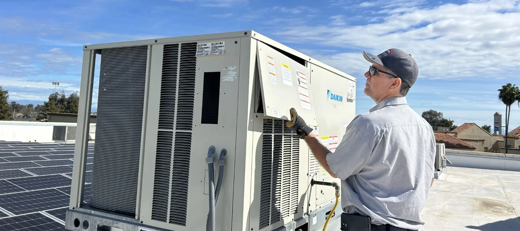 A technician in a gray uniform and cap inspects a large rooftop HVAC unit on a sunny day, with solar panels and palm trees visible in the background.