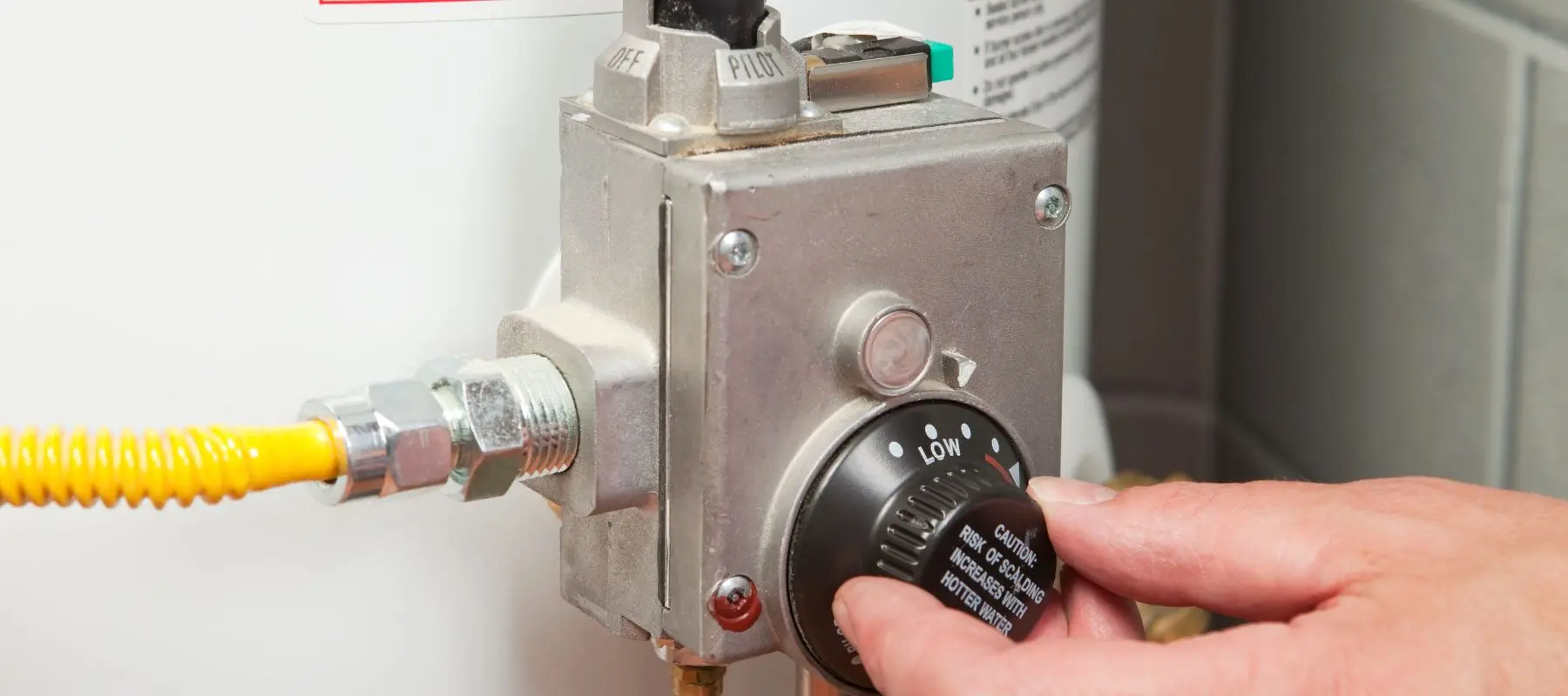 A hand adjusts the temperature dial on a water heater, turning the knob from low to high. The heaters control panel and connected pipes are visible.