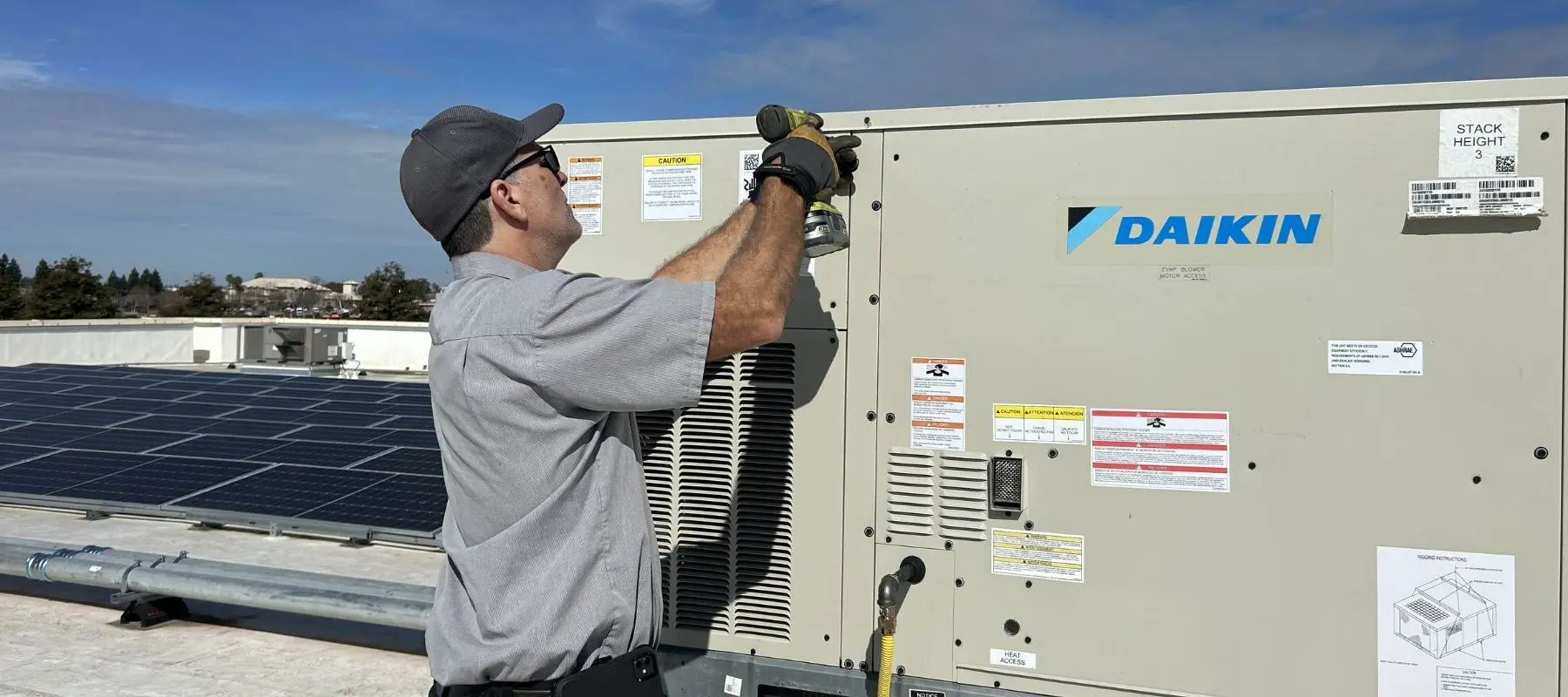 A man in work attire uses a power tool to service a Daikin HVAC unit on a rooftop next to solar panels under a partly cloudy sky.