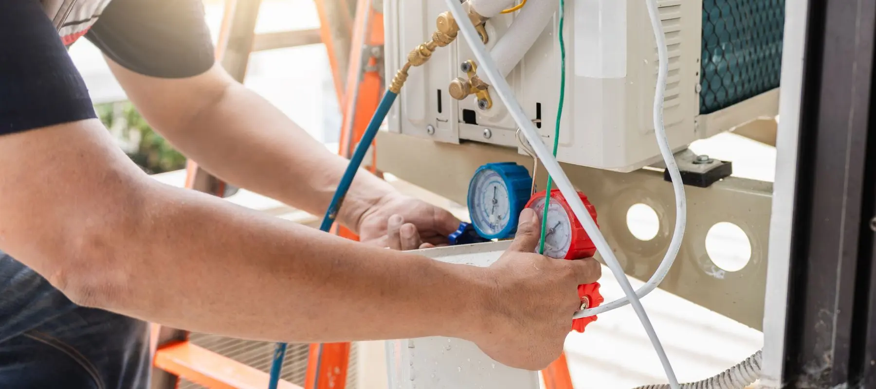 A person uses a gauge to check or repair an air conditioning unit, adjusting hoses connected to the device while standing on a ladder.