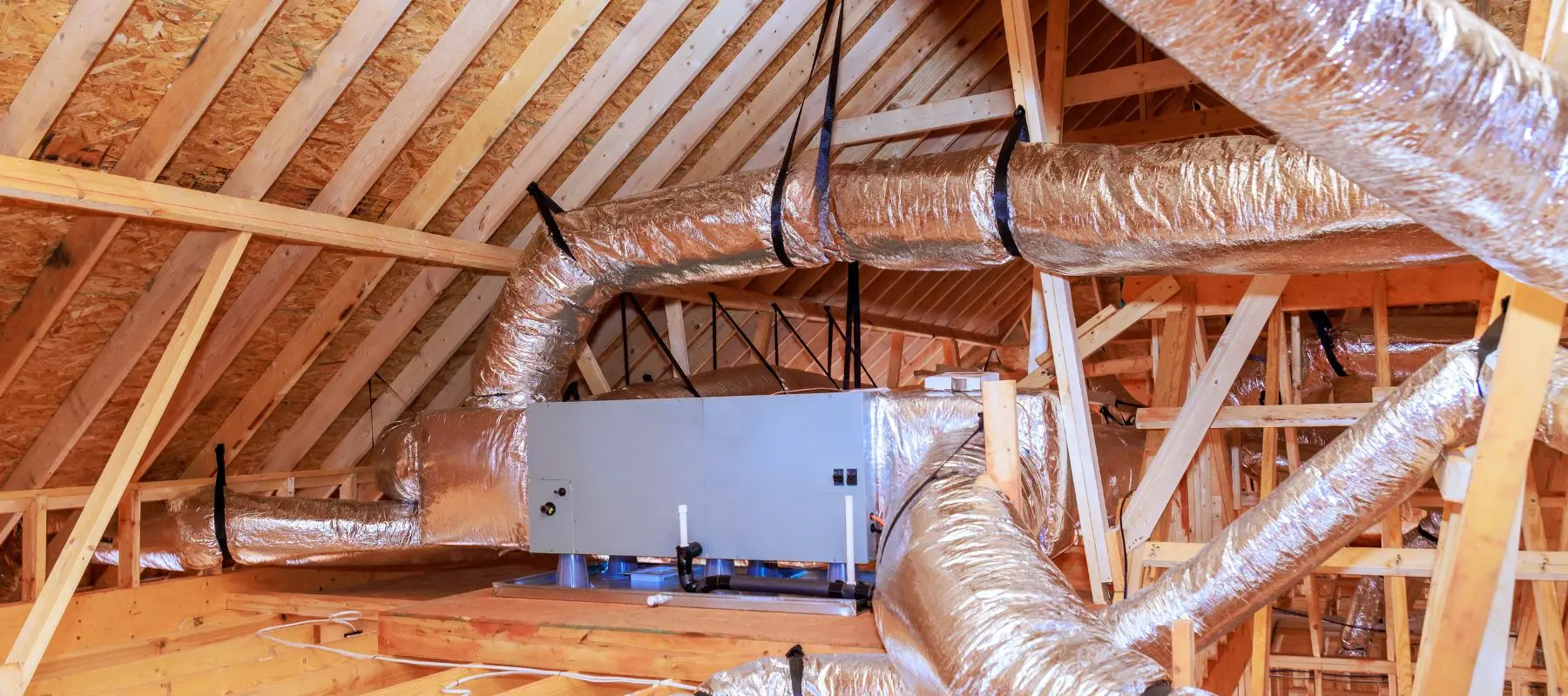 HVAC ductwork system with silver insulated pipes and a central unit installed in an unfinished attic with wooden beams and exposed roof framing.