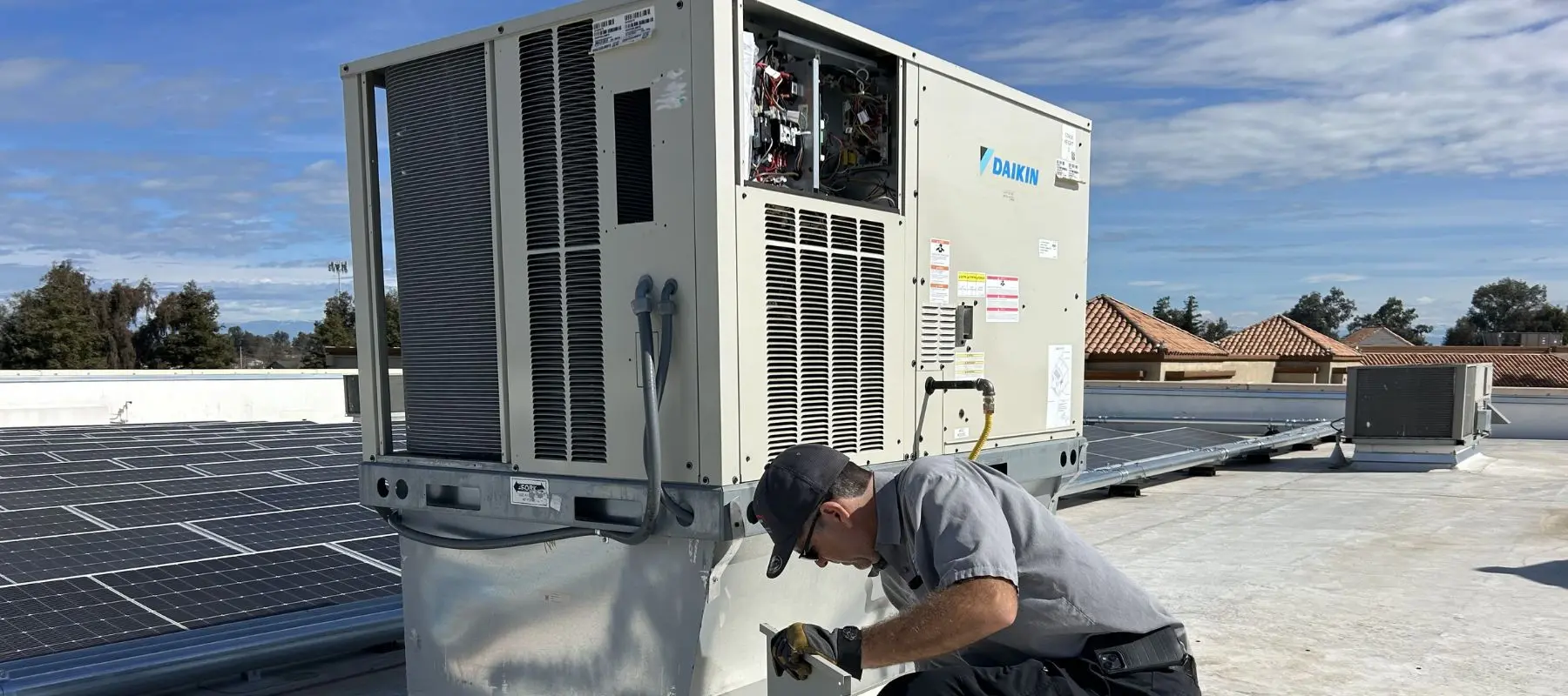 A technician in uniform works on an HVAC unit on a rooftop near solar panels, with houses and trees visible in the background under a partly cloudy sky.