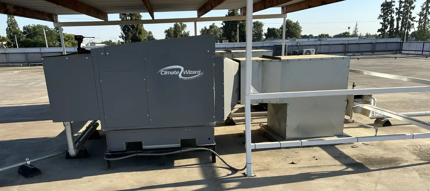 Large HVAC units labeled Climate Wizard are installed on a flat rooftop, surrounded by metal pipes and beams, under a partial roof. Trees and a fence are visible in the background under a clear sky.