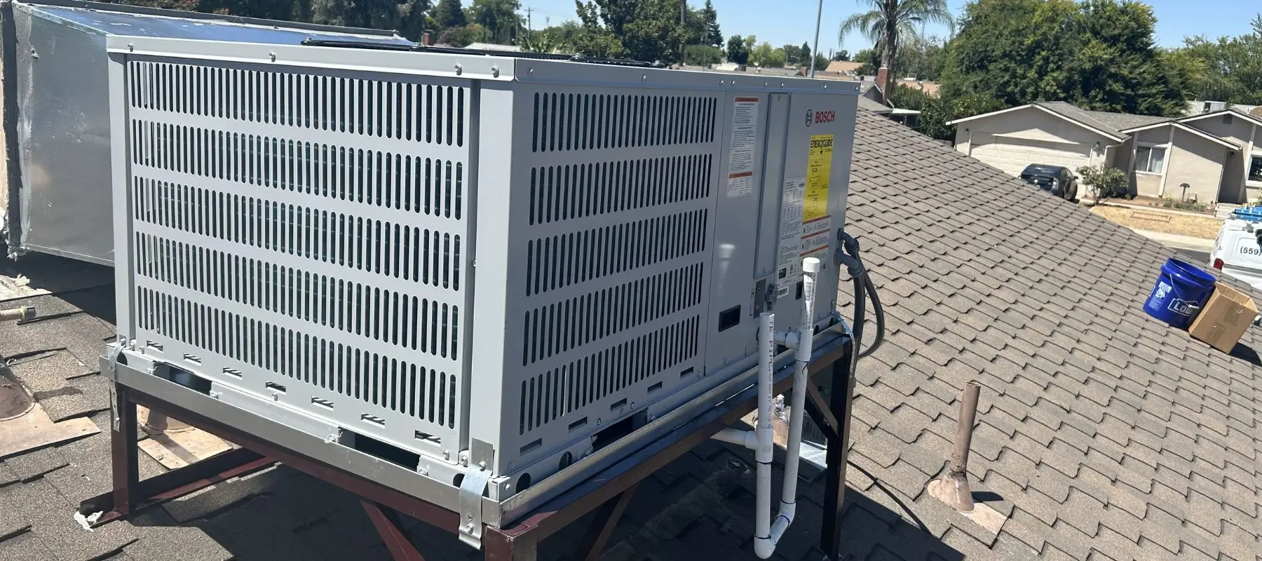 A large metal HVAC unit is installed on a shingled rooftop with pipes and cables connected, surrounded by trees and nearby houses under a clear sky.