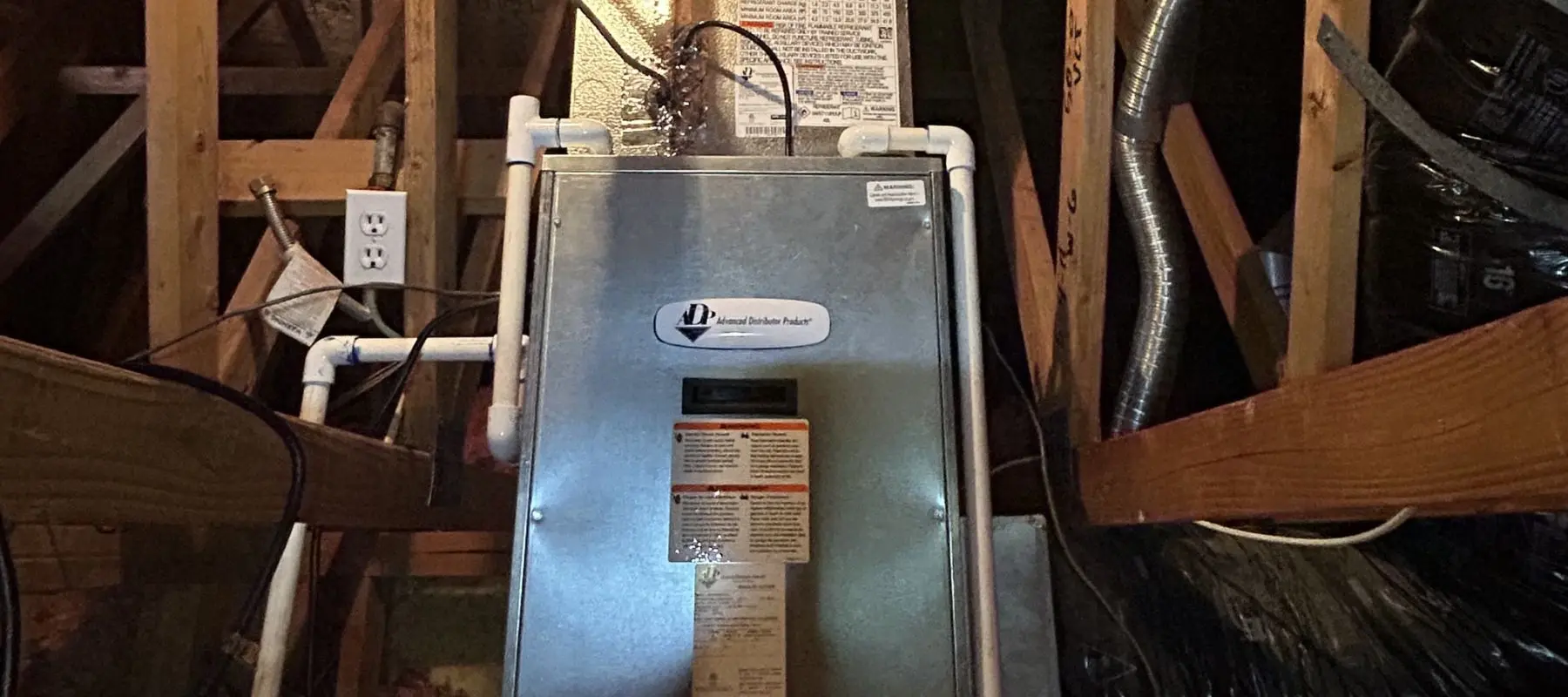 A central air conditioner unit installed in an attic, surrounded by wooden beams, electrical outlets, PVC pipes, and metal ductwork.