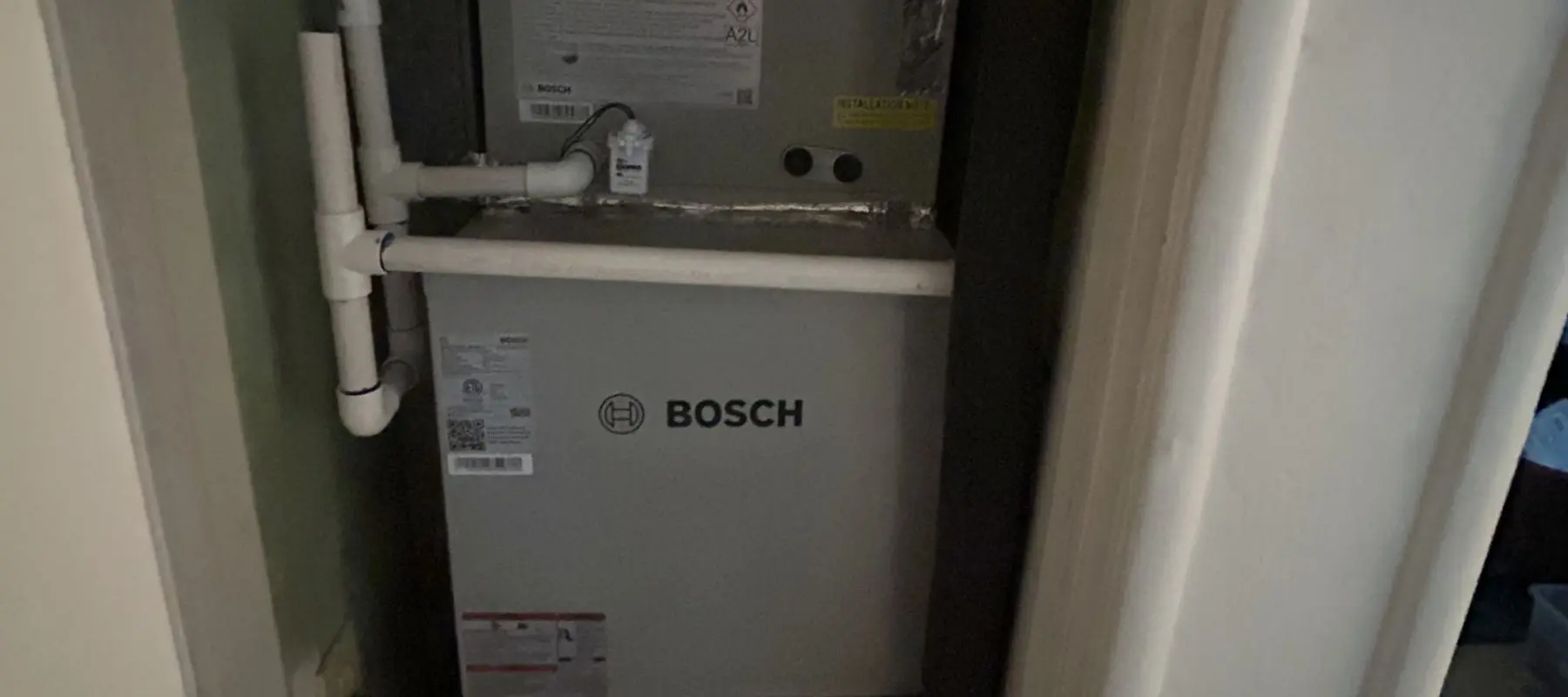 Bosch HVAC unit installed in a small closet, surrounded by white PVC pipes and partially enclosed by the doorway.