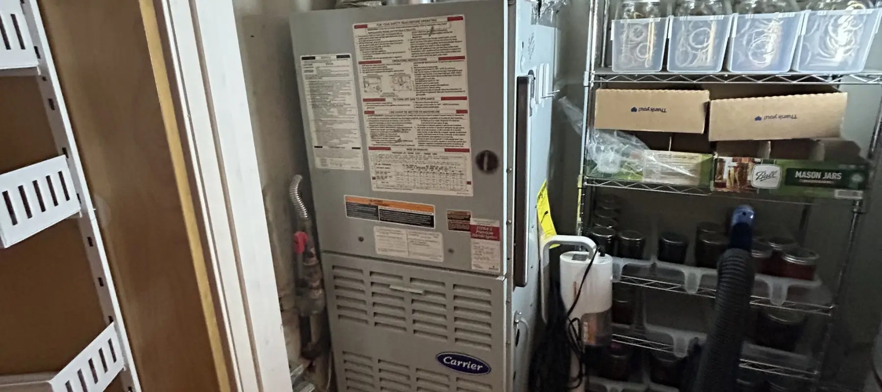 A Carrier furnace is installed in a small utility closet, with metal shelving nearby holding jars, boxes, and household items. The furnace is covered in instruction labels.