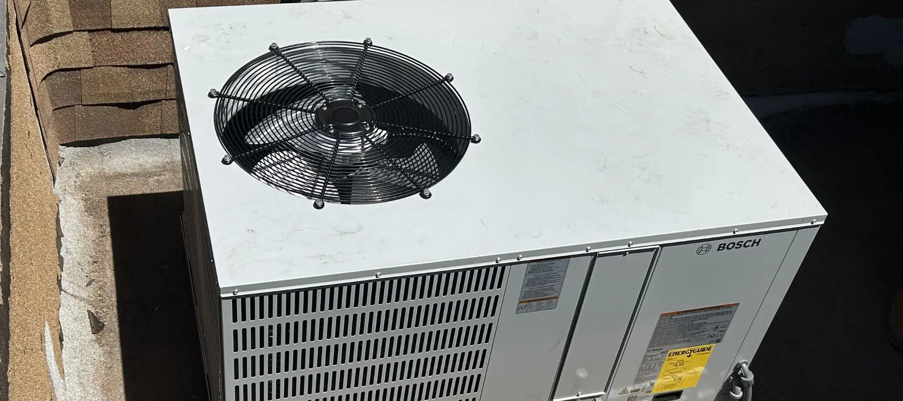 A rooftop air conditioning unit with a visible black fan on top, metal vent panels on the side, and a BOSCH logo on the front. Sunlight casts shadows over the unit.