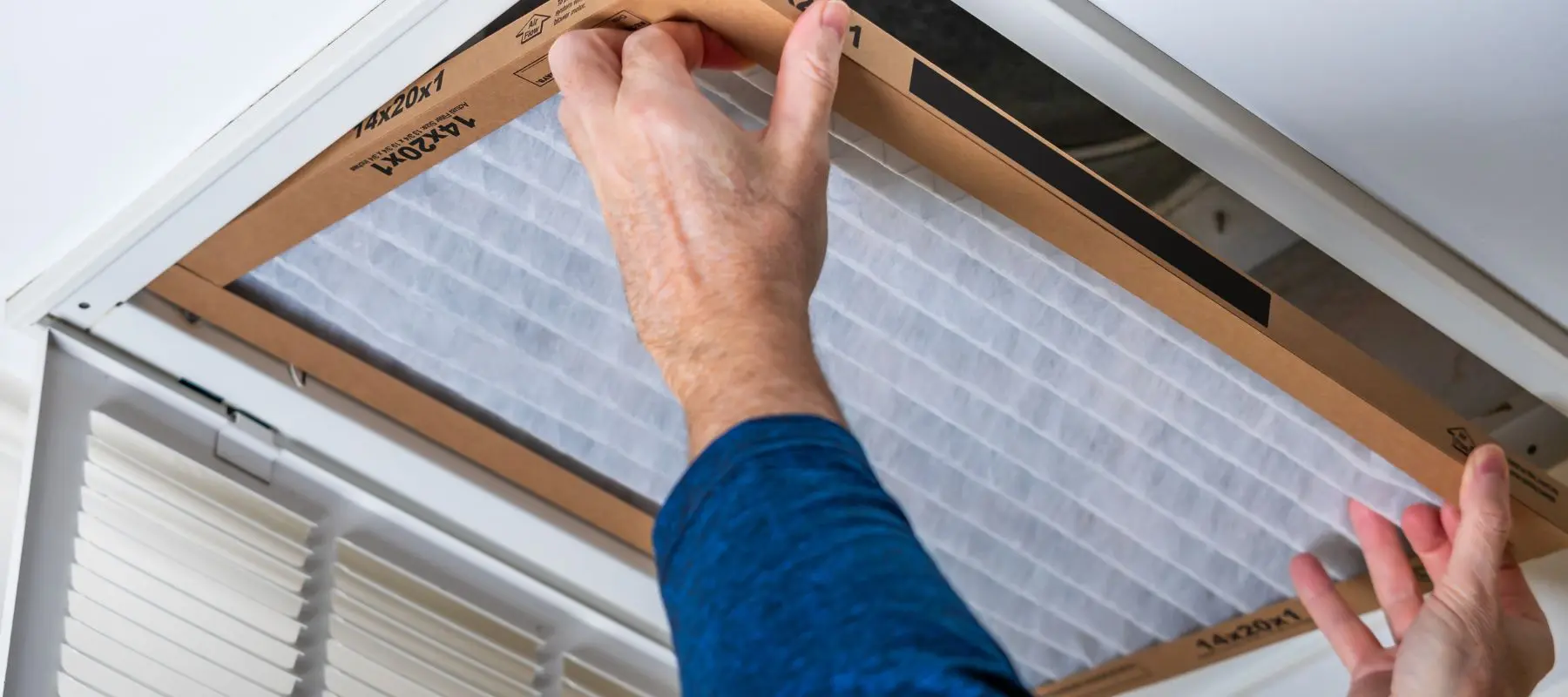 A person in a blue shirt installs or replaces an air filter in a ceiling vent, using both hands to fit the rectangular filter into place.