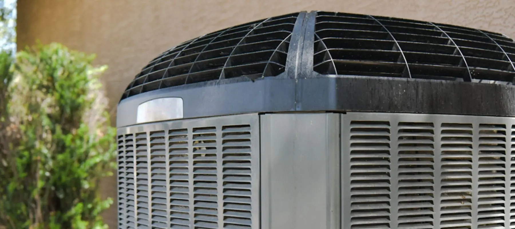 Close-up of an outdoor air conditioning unit with a metal grill and vented sides, positioned next to a beige wall and some greenery in the background.