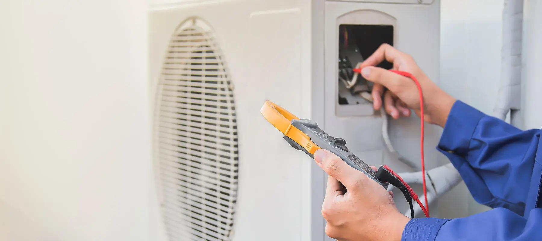 A technician in a blue uniform uses a digital multimeter to check electrical connections on an outdoor air conditioning unit.