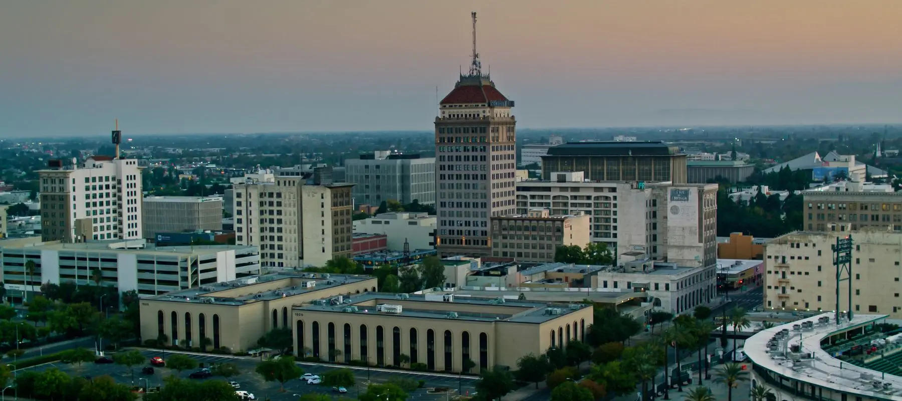 Downtown cityscape at sunset with a tall, pointed-roof building in the center, surrounded by other office buildings and streets lined with trees. The sky is a gradient of orange and blue.