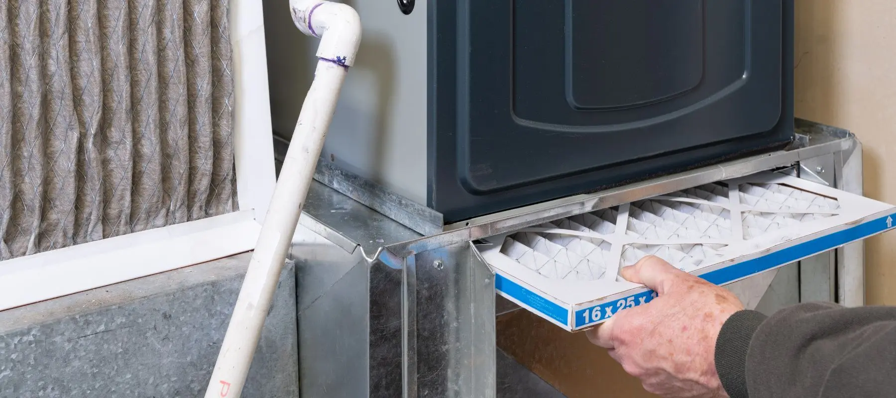 A person is sliding a new air filter into a home HVAC system, with an old dirty filter visible nearby and some PVC pipes connected to the unit.