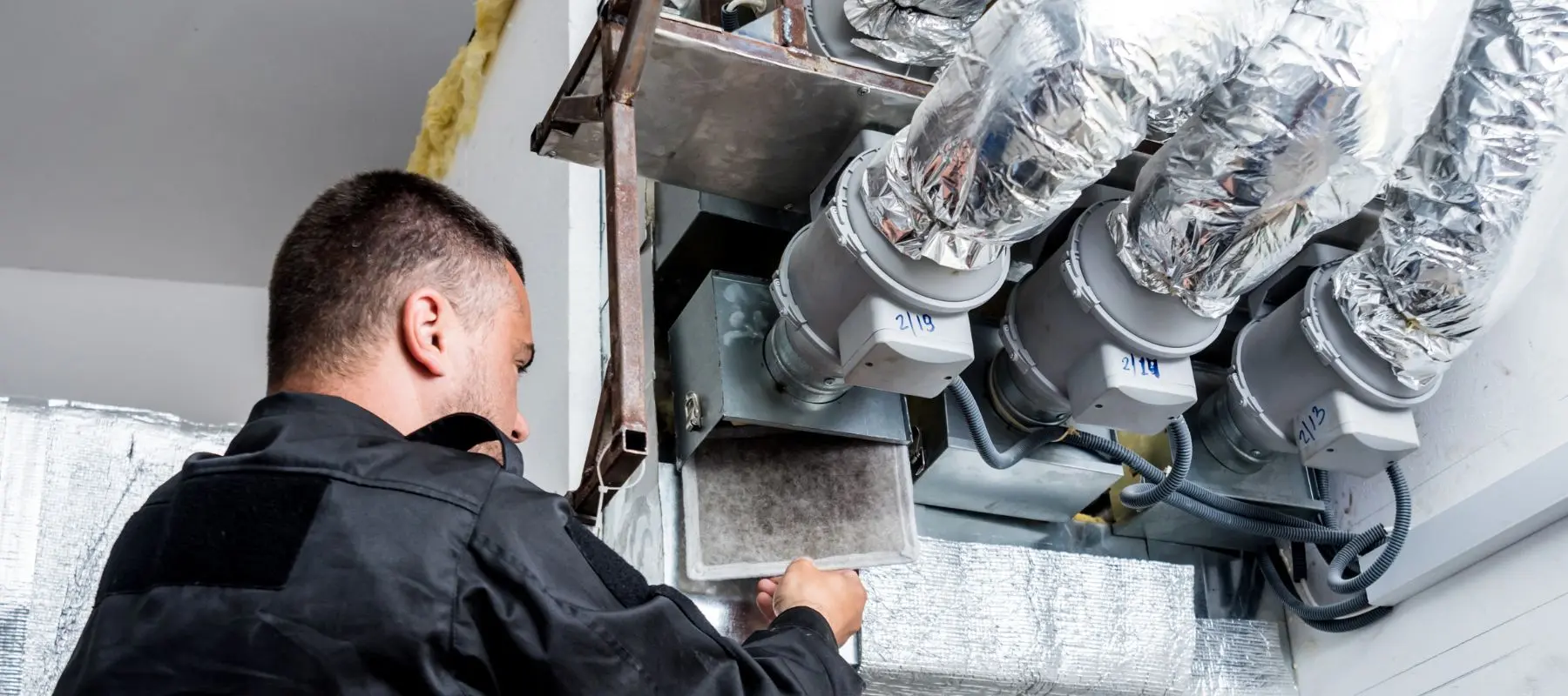A technician in a black uniform inspects and maintains HVAC ductwork and filters, surrounded by metallic pipes covered in insulation material.