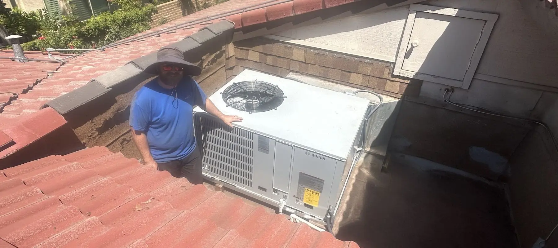 A person in a blue shirt and wide-brimmed hat stands on a tiled roof next to a large outdoor air conditioning unit next to a brick wall.