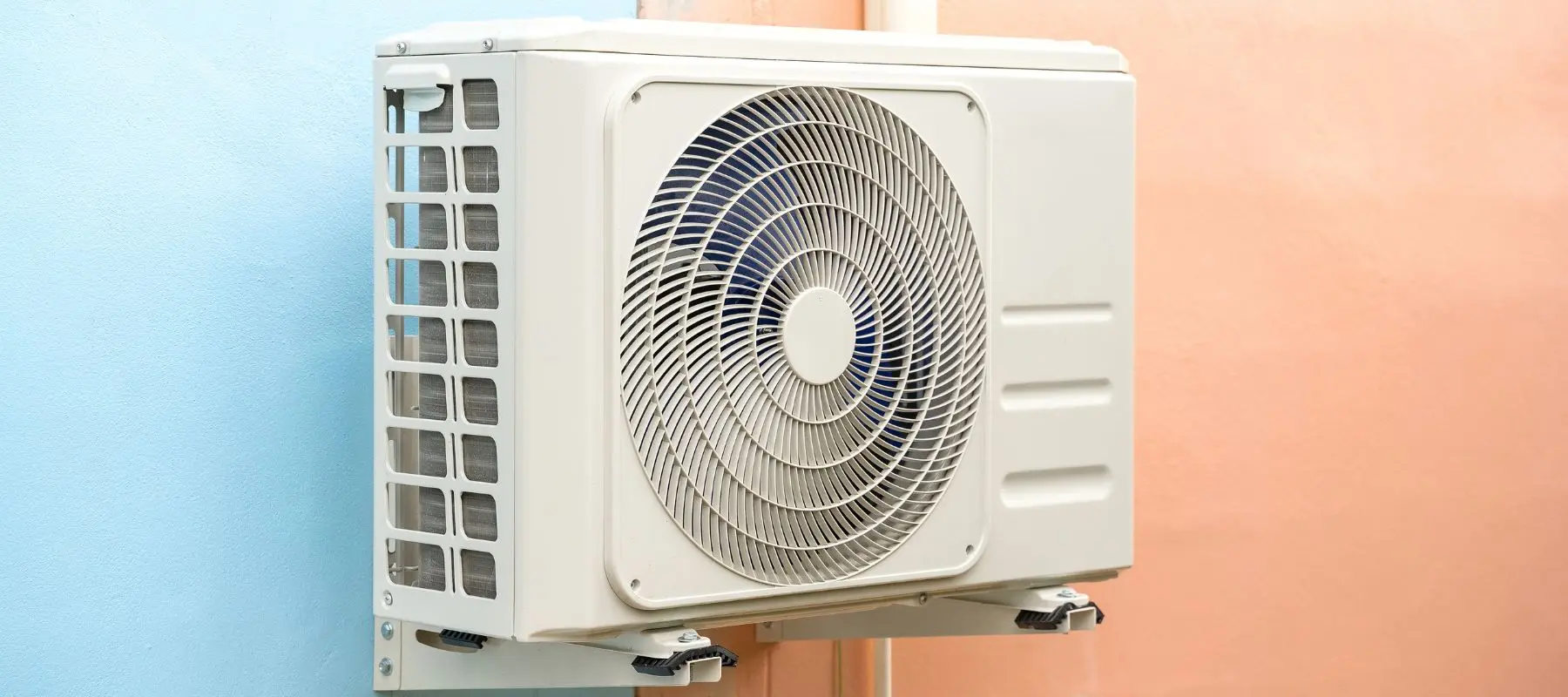 A white air conditioning unit is mounted on an outdoor wall, with a blue wall on the left and a peach wall on the right.