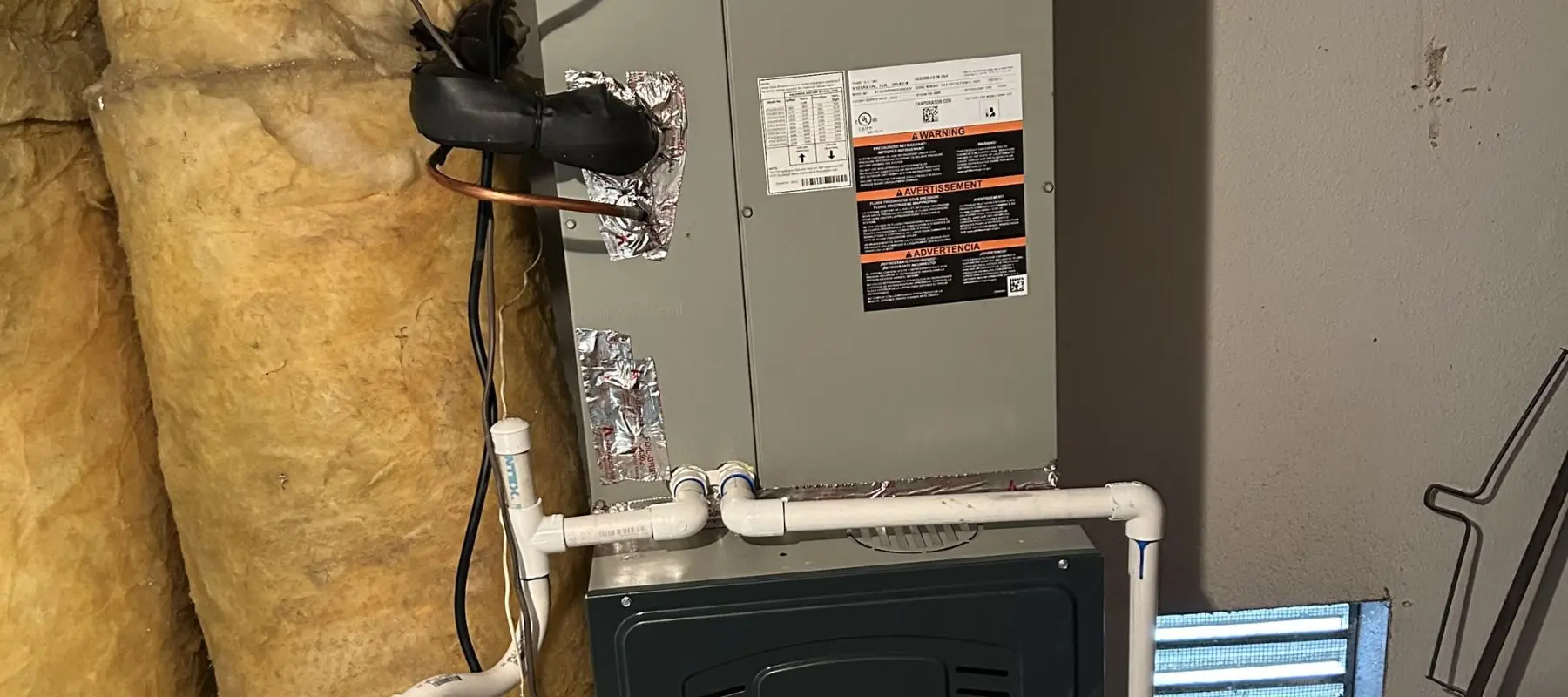 Close-up of an HVAC unit installed next to yellow insulation in a utility room, with black and white pipes connected and various warning and instruction labels visible on the unit.
