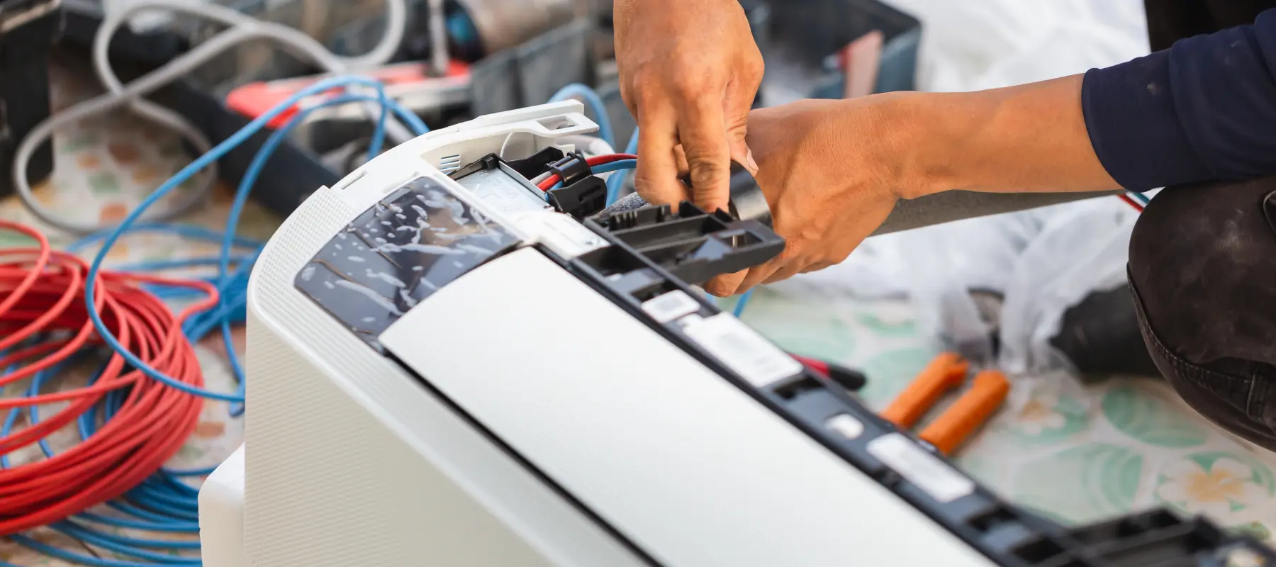 A person repairs an air conditioning unit, using both hands to adjust parts inside the open machine. Electrical tools and colorful wires are visible in the background.