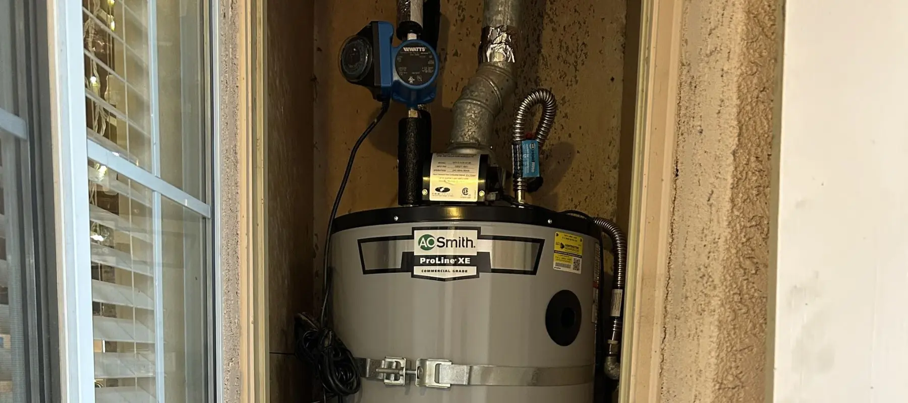 A water heater labeled AO Smith ProLine XE is installed in a small, unfinished closet. Pipes, ducts, and a blue pump are connected above and around the unit. Light enters from a window on the left side.