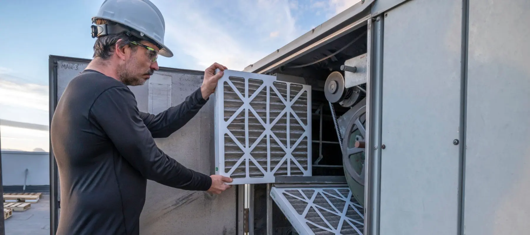 A worker in safety gear replaces an air filter in a large HVAC unit outdoors, ensuring proper maintenance and airflow.