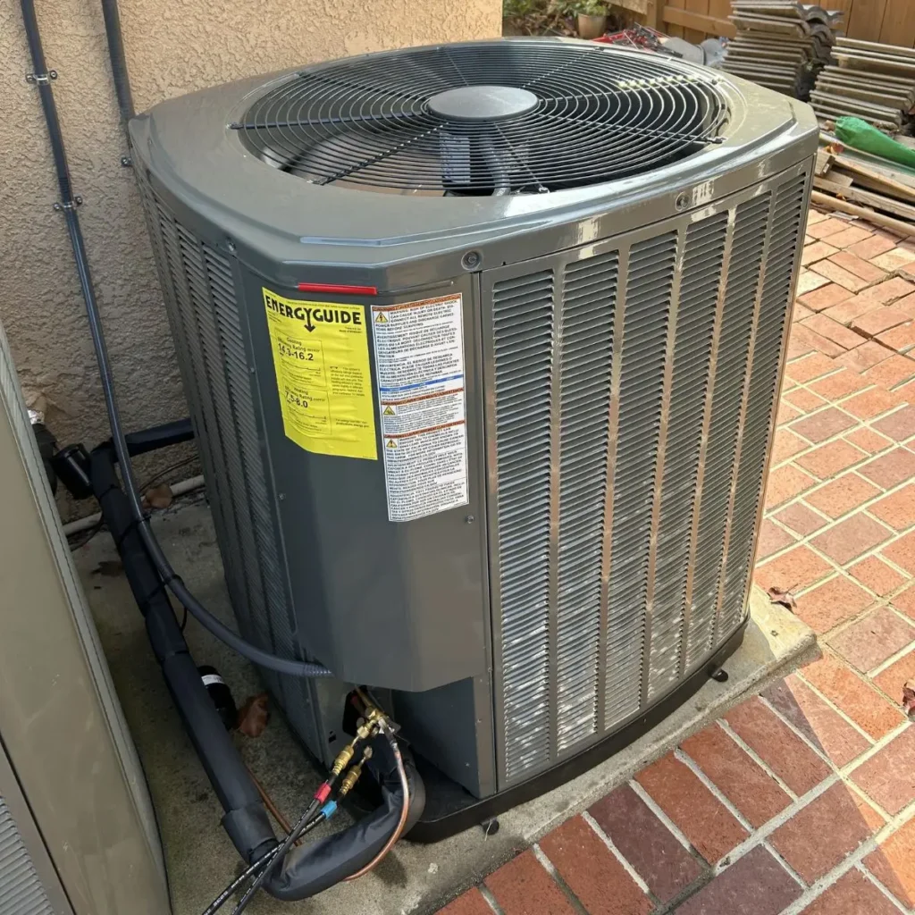 Outdoor central air conditioning unit on a concrete slab next to a brick patio, with an EnergyGuide label and refrigerant lines connected at the base.